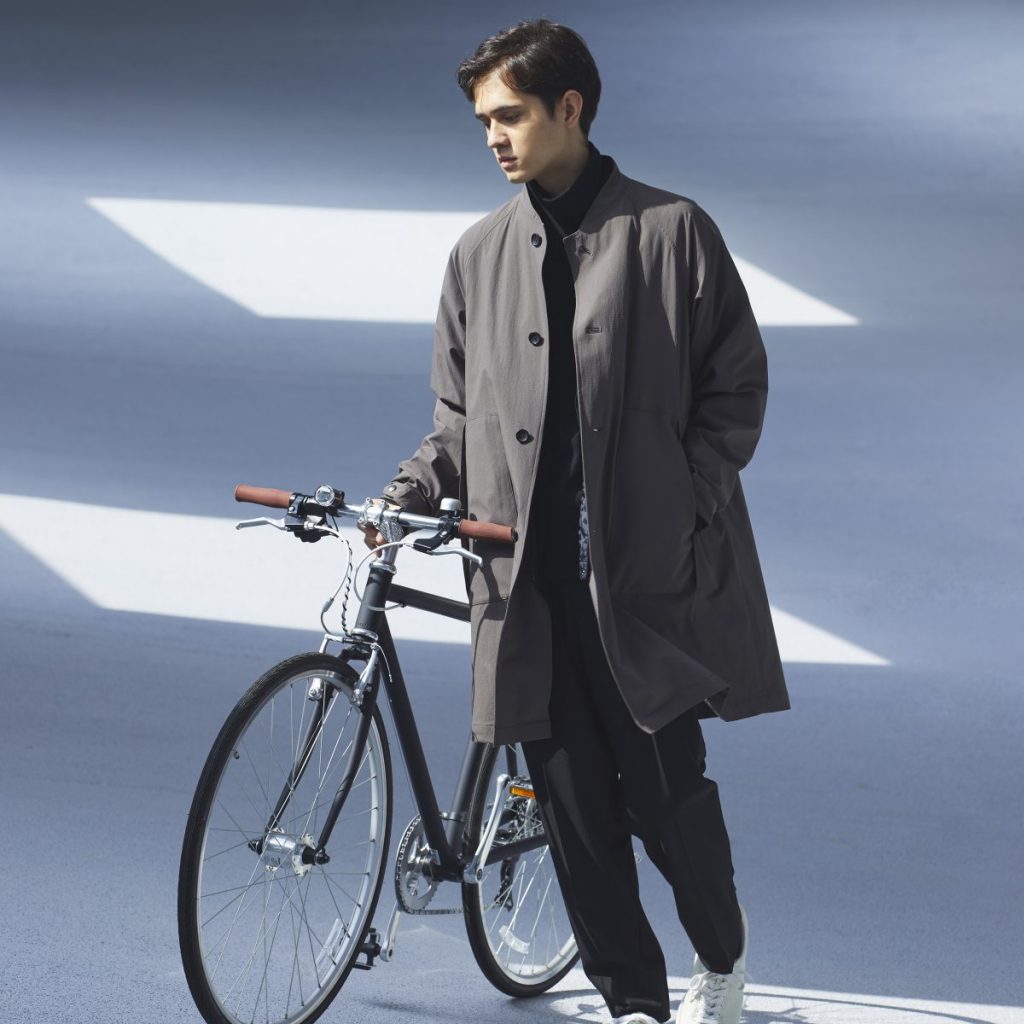 1人の男性が自転車を右手で支えながら、立ち止まっている画像。黒のスタンドコート、黒っぽいパンツ、白のスニーカーのコーディネート。左手はポケット。